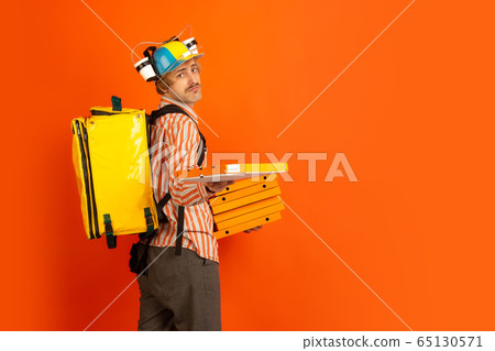 Contacless delivery service during quarantine. Man delivers food and shopping bags during insulation. Emotions of deliveryman isolated on orange background. Contacless delivery service during quarantine. Man delivers food and shopping bags during insulation. Emotions of deliveryman isolated on orange background. 65130571