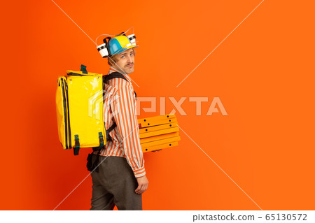 Contacless delivery service during quarantine. Man delivers food and shopping bags during insulation. Emotions of deliveryman isolated on orange background. Contacless delivery service during quarantine. Man delivers food and shopping bags during insulation. Emotions of deliveryman isolated on orange background. 65130572