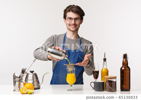 Barista, cafe worker and bartender concept. Portrait of good-looking bearded man in apron pouring drink in glass from shaker, prepare cocktail on bar counter, smiling at client 65130833