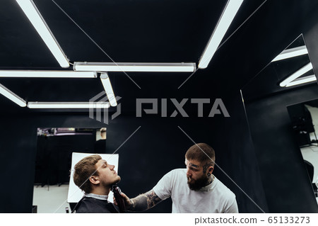 Tattooed Barber trimming bearded man with shaving machine in barbershop. Hairstyling process. Hairstylist cutting the beard of a bearded male. 65133273