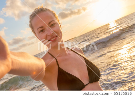 Smiling young woman taking a selfie photo at sandy beach by the sea at sunset 65135856