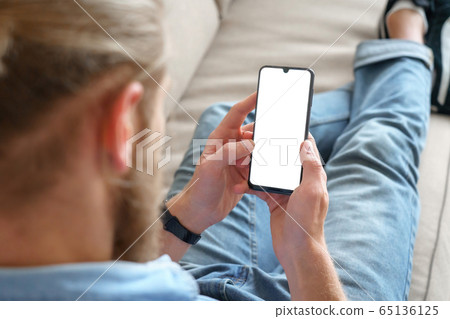 Young male tech user relaxing on sofa using smartphone mock up blank white screen. 65136125