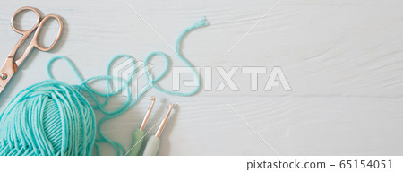 Yarn of green, turquoise, aquamarine and blue colors. White wood background. Knitting needles and crochet hooks. Scissors. White long background. 65154051
