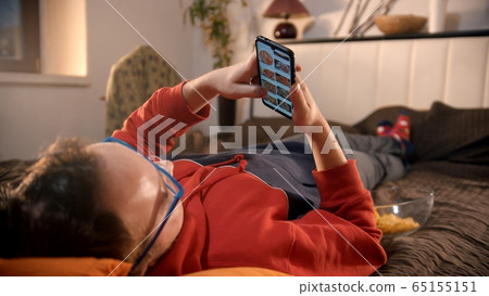 Young man lying on the bed and scrolling pictures of pizza on his phone and about to order it 65155151