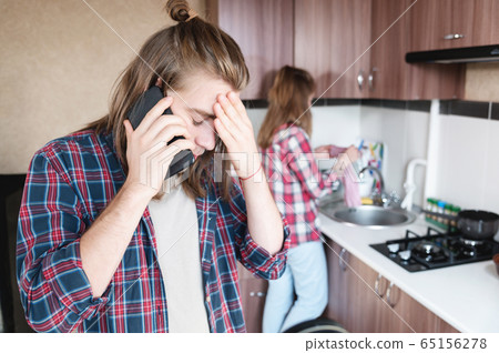 A long-haired man is upset talking on the phone or calling a food and drug delivery service home standing in the kitchen 65156278