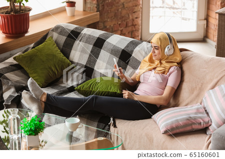 A pretty young muslim woman at home during quarantine and self-insulation, using smartphone for comunication, online lessons, shopping A pretty young muslim woman at home during quarantine and self-insulation, using smartphone for comunication, online lessons, shopping 65160601