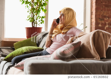 A pretty young muslim woman at home during quarantine and self-insulation, using smartphone for comunication, online lessons, shopping A pretty young muslim woman at home during quarantine and self-insulation, using smartphone for comunication, online lessons, shopping 65160602