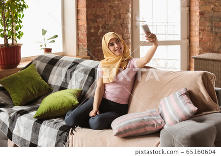 A pretty young muslim woman at home during quarantine and self-insulation, using smartphone for selfie or videocall, online lessons, shopping A pretty young muslim woman at home during quarantine and self-insulation, using smartphone for selfie or videocall, online lessons, shopping 65160604