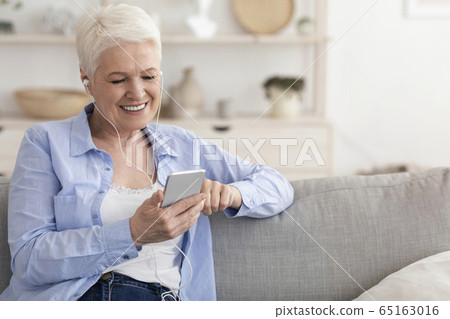 Modern Senior Woman Listening Music On Smartphone At Home, Wearing Earphones 65163016