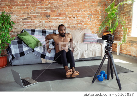 Young african-american man teaching at home online courses of fitness, aerobic, sporty lifestyle during quarantine, reording on camera, streaming 65165057