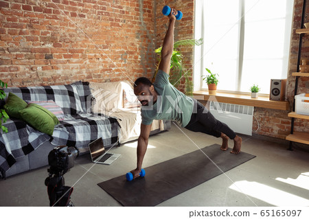 Young african-american man teaching at home online courses of fitness, aerobic, sporty lifestyle during quarantine, reording on camera, streaming 65165097
