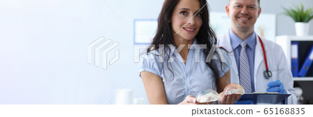 Girl in clinic holds breast implants near doctor 65168835
