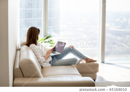 Relaxed woman using laptop sitting on sofa at luxury home 65169010