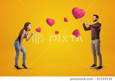 Side view of young couple on amber background playing with cute valentine hearts that float in air, man trying to catch one heart. Side view of young couple on amber background playing with cute valentine hearts that float in air, man trying to catch one heart. 65169536