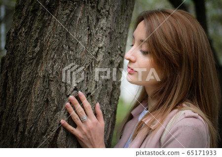 A young attractive woman with a blonde hair posing near a big tree in a spring park outdoors. She leans against a tree to touch the bark A young attractive woman with a blonde hair posing near a big tree in a spring park outdoors. She leans against a tree to touch the bark 65171933