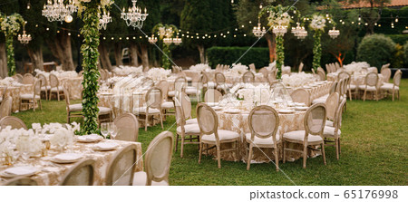 Wedding dinner table reception. Elegant tables for guests with cream tablecloths with patterns, on green lawn, with garlands and chandeliers hanging over them. Chairs with round back 65176998