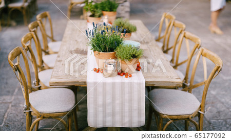 Wedding dinner table reception at sunset outside. Ancient rectangular wooden tables with rag runner, wooden vintage chairs, lavender pots, cherry tomatoes and clay pots with lemons on tables 65177092