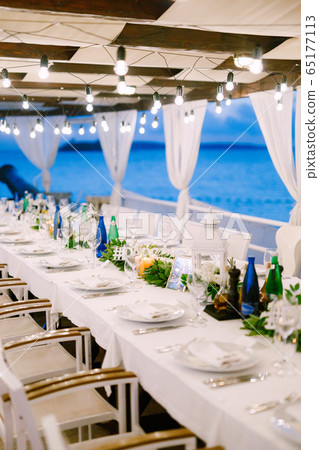 Wedding dinner table reception. A long table, against the sea, with a white tablecloth, a garland with incandescent light bulbs over it. Wooden white chairs. Flower composition in the center of table 65177113