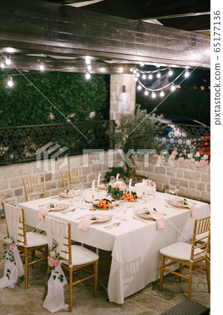 Wedding dinner table reception. Rectangular table for six people with a white tablecloth and pink napkins. The golden chairofchia is decorated with fabric and pink flowers. Garland burns over table.  65177136