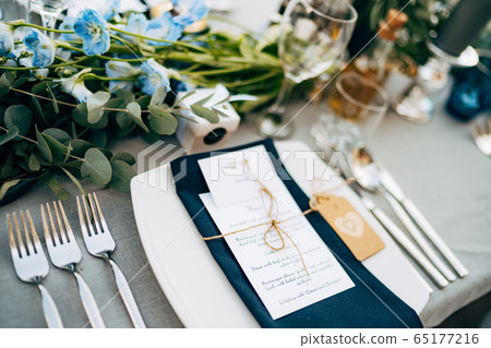 Wedding dinner table reception. A square plate with a blue cloth towel, knives and forks next to the plate. Flower composition with eucalyptus leaves in the center of the table and burning candles. 65177216