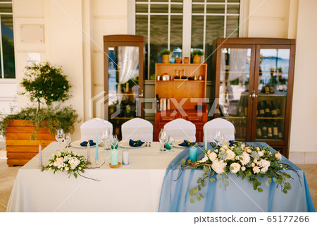 Wedding dinner table reception. White rectangular table of newlyweds with four chairs, white-blue tablecloth, floral arrangements with eucalyptus leaves, against background of wine cabinets 65177266