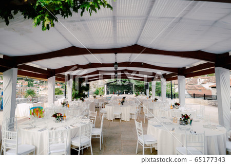 Wedding dinner table reception. Round tables with white tablecloths and white Chiavary chairs under a large white tent, against the backdrop of a musical group. 65177343
