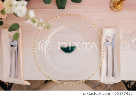 Wedding dinner table reception. Close-up of wildcard with gold beads, transparent glass. Runner of pink silk. Candles in golden candlesticks and flowers in the center of the table.  65177433