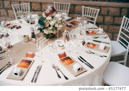 Wedding dinner table reception. Round table with white tablecloth with brown runner on table. Vienna nested in brown napkins, a bouquet of flowers is on the table. White Chiavari chairs with pillows. Wedding dinner table reception. Round table with white tablecloth with brown runner on table. Vienna nested in brown napkins, a bouquet of flowers is on the table. White Chiavari chairs with pillows. 65177477