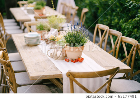 Wedding dinner table reception at sunset outside. Ancient rectangular wooden tables with rag runner, wooden vintage chairs, lavender pots, cherry tomatoes and clay pots with lemons on tables 65177491