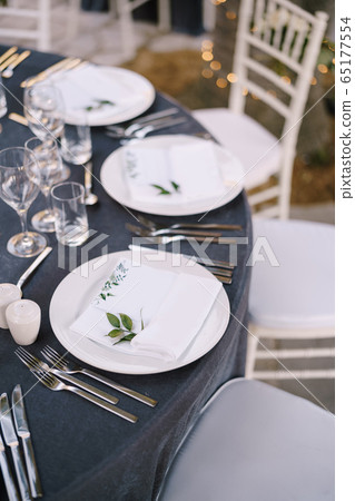 Wedding dinner table reception. White round plates on a round table with gray tablecloth, white Chiavari chairs with white pillows. A floral arrangement in the center of the table. 65177554