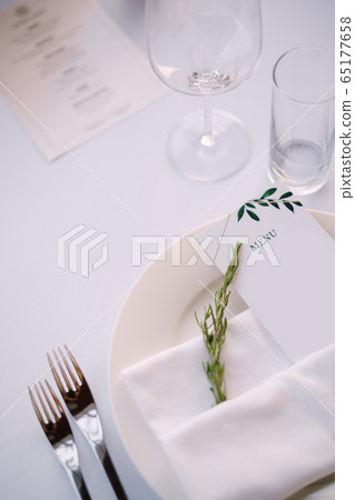 Wedding dinner table reception. White plate with cloth towel with paper menu and sprig of rosemary, against the background of white tablecloth 65177658