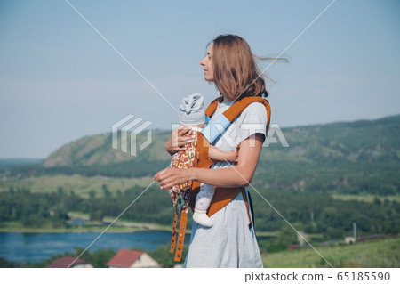 a woman with a child in a special carrier, a kangaroo in a walk. Village houses, the forest and river as the background. The concept of summer, warmth, freedom, village life, sunburn, childhood 65185590