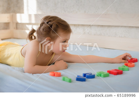 girl 3 years Caucasian in yellow dress lying on wooden bed on blue bedspread and learning in play form numbers and count, concept of home education 65189996