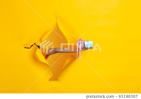 Female hand giving gift in torn yellow paper hole background, celebration 65190307