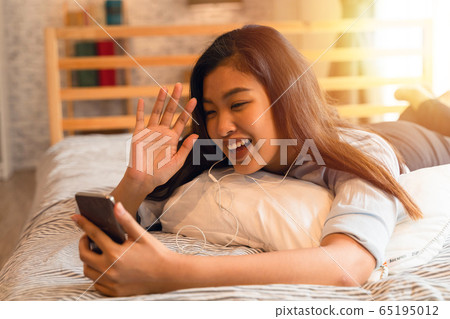 Portrait of happy young Asian girl in casual clothing lying down on bed while making a video call with smartphone and waving in bedroom at home. Video conferencing technology concept Portrait of happy young Asian girl in casual clothing lying down on bed while making a video call with smartphone and waving in bedroom at home. Video conferencing technology concept 65195012