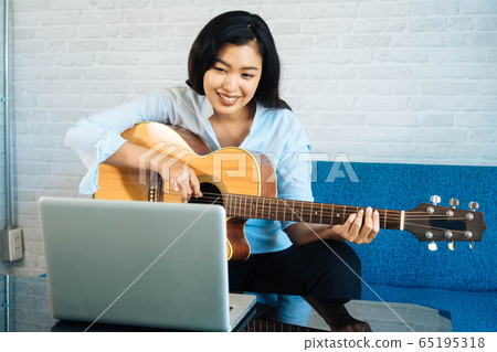 Young Asian woman practicing and learning how to play guitar on laptop computer monitor. Female guitarist watching online tutorial 65195318