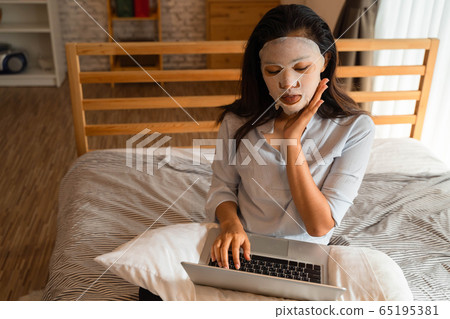Portrait of young Asian girl applying facial mask on her face while using computer laptop in bedroom. Woman's Beauty and Facial care concept 65195381