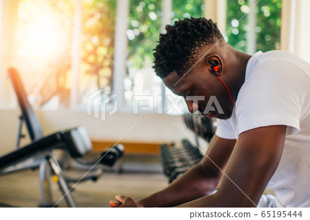 Side view of tired African American man in sportswear listening to music in wireless earphones while sitting on bench and resting during training in gym 65195444