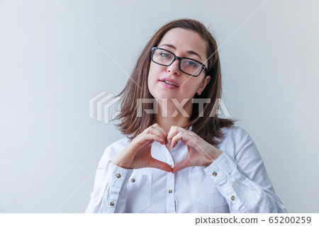 Beautiful happy girl smiling. Beauty simple portrait young woman in eyeglasses showing heart sign with hands isolated on white background. Positive human emotion facial expression body language. Beautiful happy girl smiling. Beauty simple portrait young woman in eyeglasses showing heart sign with hands isolated on white background. Positive human emotion facial expression body language. 65200259