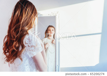 Beauty makeup morning rutine love yourself concept. Young teenage girl looking at reflection in mirror. Young positive woman wearing white dress posing in bright light room against white wall. Beauty makeup morning rutine love yourself concept. Young teenage girl looking at reflection in mirror. Young positive woman wearing white dress posing in bright light room against white wall. 65200274