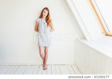 Happy teenage girl smiling. Portrait young happy positive teen woman in grey dress at home in bright room against white wall. European woman. Positive human emotion facial expression body language. 65200277