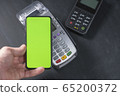 Top view of person using contactless payment with smartphone. Clear green mockup screen template of phone and pos terminal on dark wooden desk with side copy space 65200372