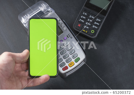 Top view of person using contactless payment with smartphone. Clear green mockup screen template of phone and pos terminal on dark wooden desk with side copy space 65200372