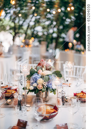 Wedding dinner table reception. Bouquet in a vase on a table of pink and white roses and blue hydrangea, with the number of the table on the plate. Burning candles in high glass candlesticks. Wedding dinner table reception. Bouquet in a vase on a table of pink and white roses and blue hydrangea, with the number of the table on the plate. Burning candles in high glass candlesticks. 65204030