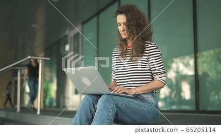 Attractive young woman with long curly hair wearing striped shirt is sitting on stairs of building Attractive young woman with long curly hair wearing striped shirt is sitting on stairs of building 65205114
