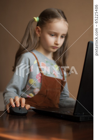 Serious little girl is concentrated in front of her black laptop while focused on doing homework during the quarantine because of the pandemia of Coronavirus Covid-19. Remote education Serious little girl is concentrated in front of her black laptop while focused on doing homework during the quarantine because of the pandemia of Coronavirus Covid-19. Remote education 65225486