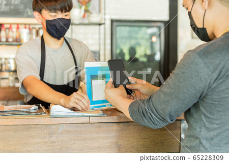 Male customer with protective mask paying bill by cell phone in cafe. Male customer with protective mask paying bill by cell phone in cafe. 65228309
