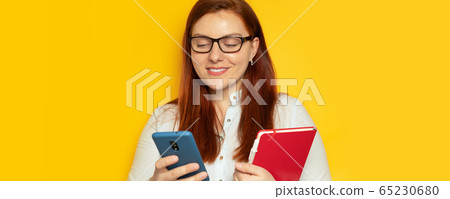 Business woman in glasses and a blouse uses a smartphone and notepad on a yellow wall background. Stay at home, work at home Business woman in glasses and a blouse uses a smartphone and notepad on a yellow wall background. Stay at home, work at home 65230680
