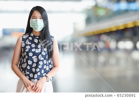 Beautiful woman wearing a medical mask and standing over blurred international airport. Beautiful woman wearing a medical mask and standing over blurred international airport. 65230891