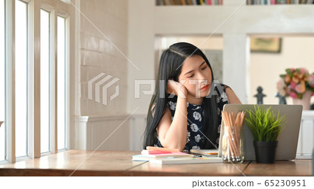 Beautiful girl keep hand on chin and looking at computer laptop while sitting at the wooden table. Beautiful girl keep hand on chin and looking at computer laptop while sitting at the wooden table. 65230951